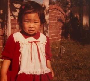 The author as a 2-3 year old girl in a red dress with a white apron looking at the camera
