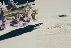 photo of purple flowers and a fountain pen laying across sheets of paper with writing on them