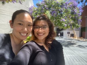 Author (Asian American woman) smiling with sister (Asian American woman) smiling, both wearing dark tops with a jacaranda tree in the background