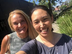 Selfie of the author (Asian American woman in a grey t-shirt) with a friend (blond white woman in grey dress) 