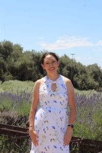 Photo of an Asian American woman (the author) in a white sleeveless dress with blue and yellow flowers in a field of lavender