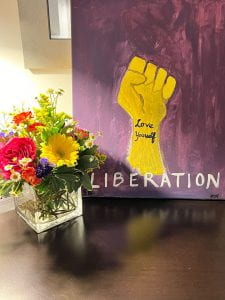 Photograph of flowers in a square vase in front of a picture of a raised fist with the words "love yourself" written on the wrist and the word liberation written below the raised fist