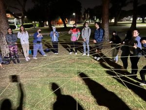 Individuals standing in a semi-circle connected by intertwined yarn