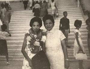 Black and white photo of two Asian women, the younger one with many flower leis around her neck, the older one in a white qipao dress 