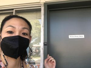 The author (an Asian American woman) wearing a Black KN95 mask standing in front of her office door pointing at it