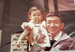 Photograph of an Chinese man holding a small Taiwanese American little girl who is his daughter, in front of a church. She is a baby with a pacifier and a white dress with a red checkered kangaroo on it. He is wearing a suit.
