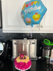 Picture of a "cake" made of flowers with six candles and a blue mylar balloon with the words happy birthday on it floating about the cake