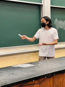 17 year old son of the author standing at a green chalkboard (in a lecture hall) with a piece of white chalk